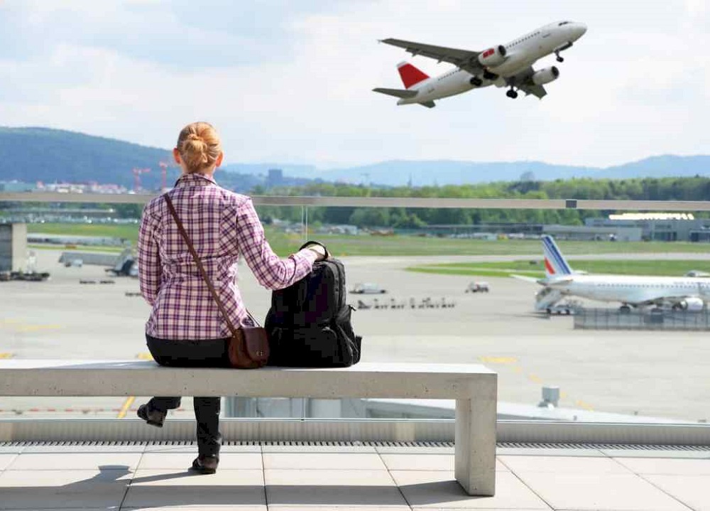 девушка в аэропорту сидит с чемоданом в зале ожидания и смотрит на улетающий самолет - авиалайнер взлетает вид из окна